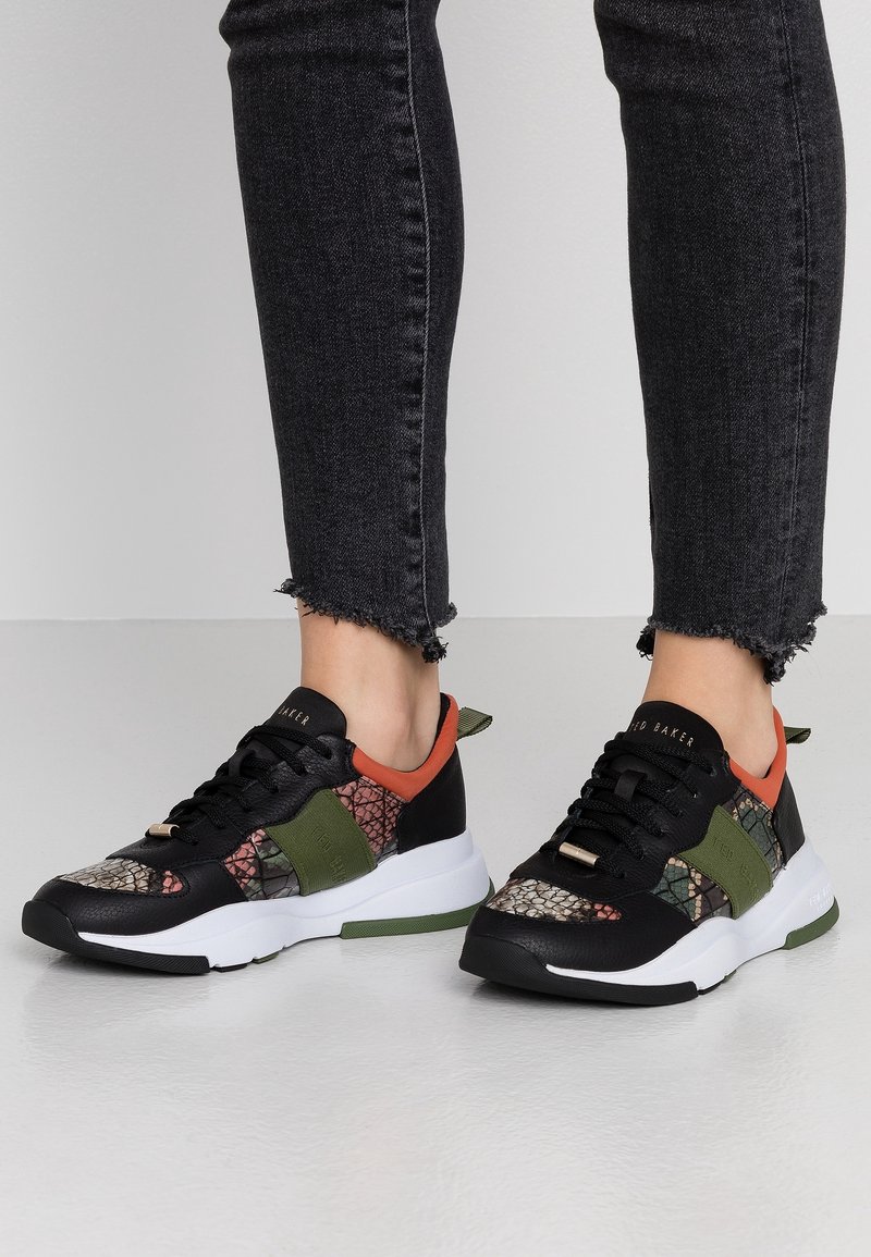 Personne portant des baskets à motif noir, vert et orange avec des semelles blanches et un jean slim noir à ourlets effilochés.