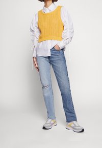 Gul stickad crop top, vit button-up skjorta, förstörda ljusblå jeans och grå sneakers med gula accenter.