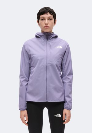 Persoon draagt een lichtpaarse capuchonvest met rits en zwarte legging, beide met The North Face-logo's, staand tegen een effen achtergrond.