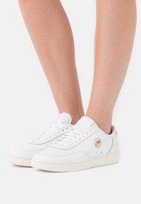 Vita lädersneakers med perforerade detaljer, platta snören och en cirkulär gul logotyp. Sulan är ljus och något texturerad.