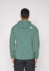 Chaqueta con capucha verde hecha de un material ligero y suave. Presenta un pequeño logo en la parte posterior y un diseño ajustado. Ideal para uso al aire libre.