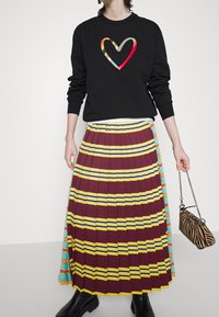 Personne portant un sweat-shirt noir avec un cœur multicolore, une jupe plissée avec des rayures bordeaux, jaunes et bleues, tenant un petit sac imprimé zèbre.