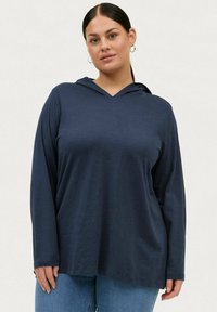 Sudadera de manga larga en color navy hecha de una tela suave, con escote en forma de V, corte relajado y capucha forrada. Combinada con jeans azules.