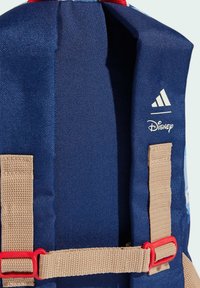 Mochila azul marino con correas beige y herrajes rojos. Presenta los logotipos de Adidas y Disney impresos en el lateral. Diseño de tela texturizada.