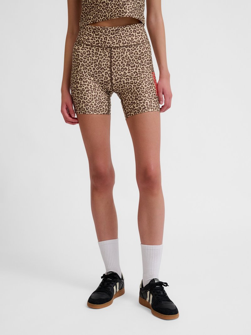 Kvinde model iført leopardprintet tætsiddende shorts, hvide crew sokker og sorte sneakers med hvide striber, stående på ensartet grå baggrund.