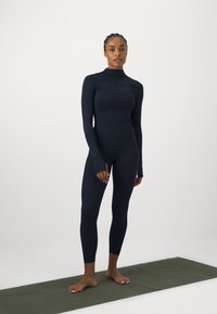 Top a maniche lunghe blu navy e leggings in tessuto elasticizzato, con collo alto e aperture per i pollici, abbinati a un tappetino da yoga verde.