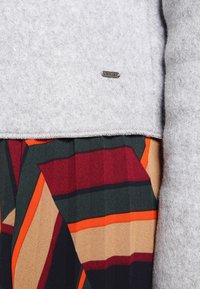 Pull en feutre gris avec une étiquette en métal argenté, superposé à une jupe plissée multicolore présentant des rayures géométriques rouges, vertes et orange.