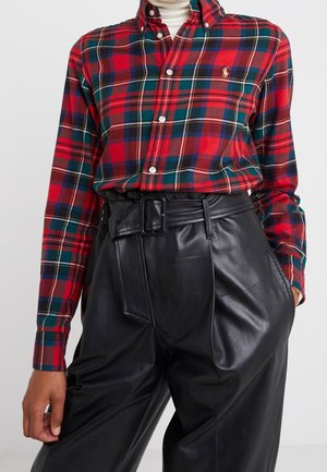Chemise à carreaux rouge, verte et bleue avec boutons, rentrée dans un pantalon en cuir noir taille haute ceinturé, les mains détendues le long du corps.