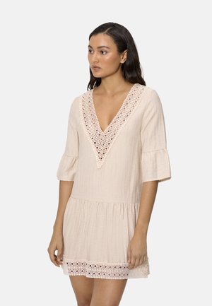 Mujer de cabello oscuro que lleva un vestido color crema con escote en V, ribete de encaje y mangas campana de tres cuartos, de pie frente a un fondo liso.