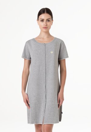 Donna che indossa un vestito grigio con bottoni, maniche corte, lunghezza al ginocchio, con un piccolo emblema a forma di cuore sul petto sinistro, in piedi davanti a uno sfondo semplice.