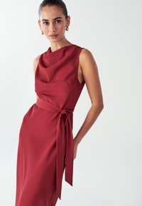 Červené šaty bez rukávů s draped výstřihem, detailem na bočním zavázání, hladkým materiálem a přiléhavou siluetou, vystavené na světlém pozadí.