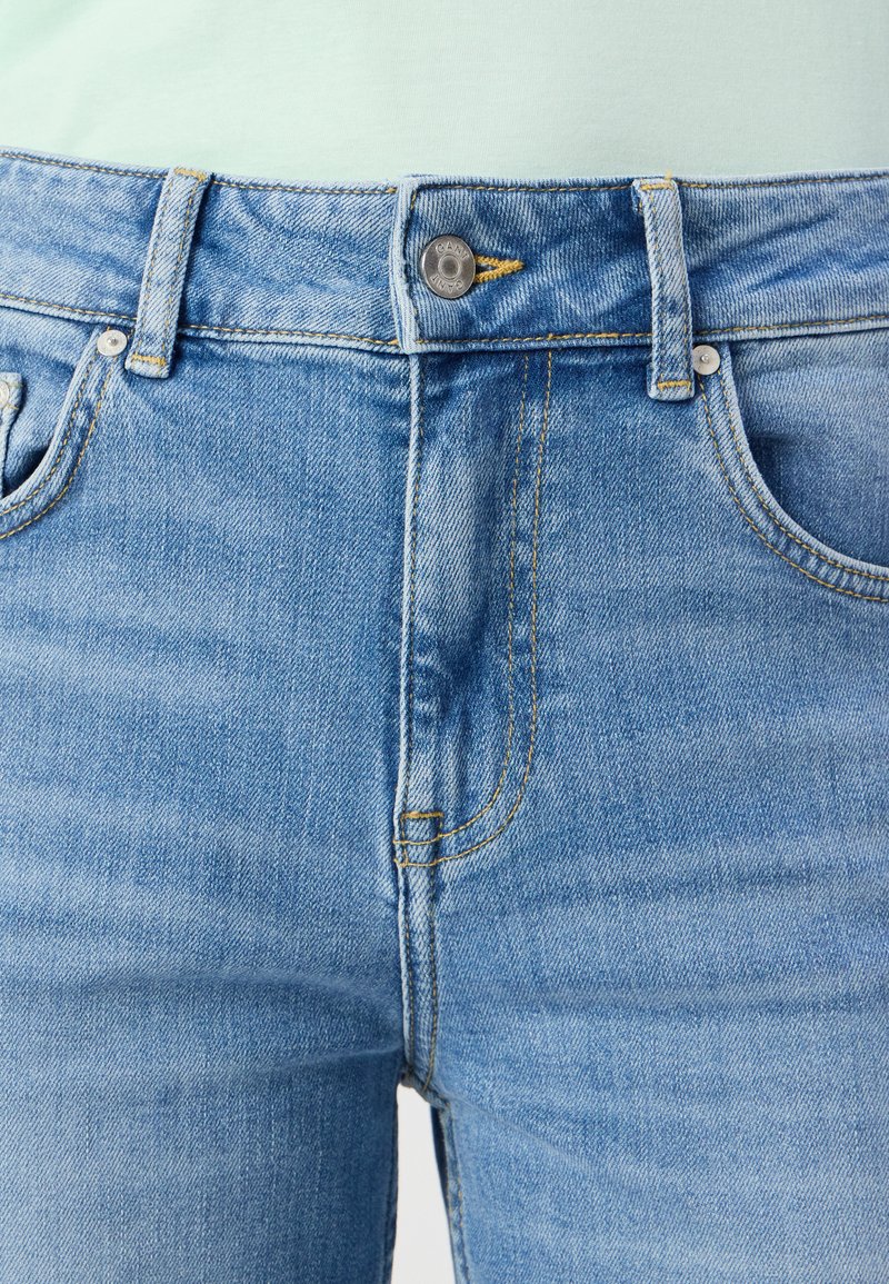 Jasnoniebieskie dżinsy mają klasyczny pięciopocketowy krój, srebrne zapięcie na guzik oraz żółte przeszycia wzdłuż szwów.