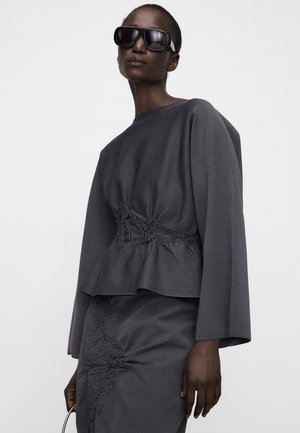 Femme portant un haut texturé gris foncé et une jupe avec un détail froncé à la taille, de grandes lunettes de soleil noires, tenant un sac à poignée fine.