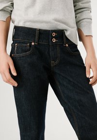 Temno modri jeans z bakrovimi dodatki, z dvojnim gumbom za zapiranje in kontrastnim oranžnim šivom. Ima sprednje žepe in naravnost krojeno hlačnico.