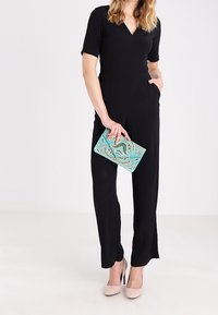 Donna che indossa una tuta nera e tacchi beige, mentre tiene una borsa a tracolla di perline turchesi su uno sfondo bianco semplice.