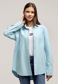 Jasnoniebieska, oversize koszula z lekkiego materiału, z guzikami z przodu, długimi rękawami i zaokrąglonym dołem. W zestawieniu z białym T-shirtem.