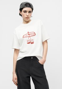 Νεαρό άτομο που φοράει λευκό μπλουζάκι με κόκκινο γραφικό καθρέφτη και ζάρια, μαύρο παντελόνι και γκρι καπέλο γυρισμένο προς τα πίσω, πόζα με το ένα χέρι στην τσέπη.