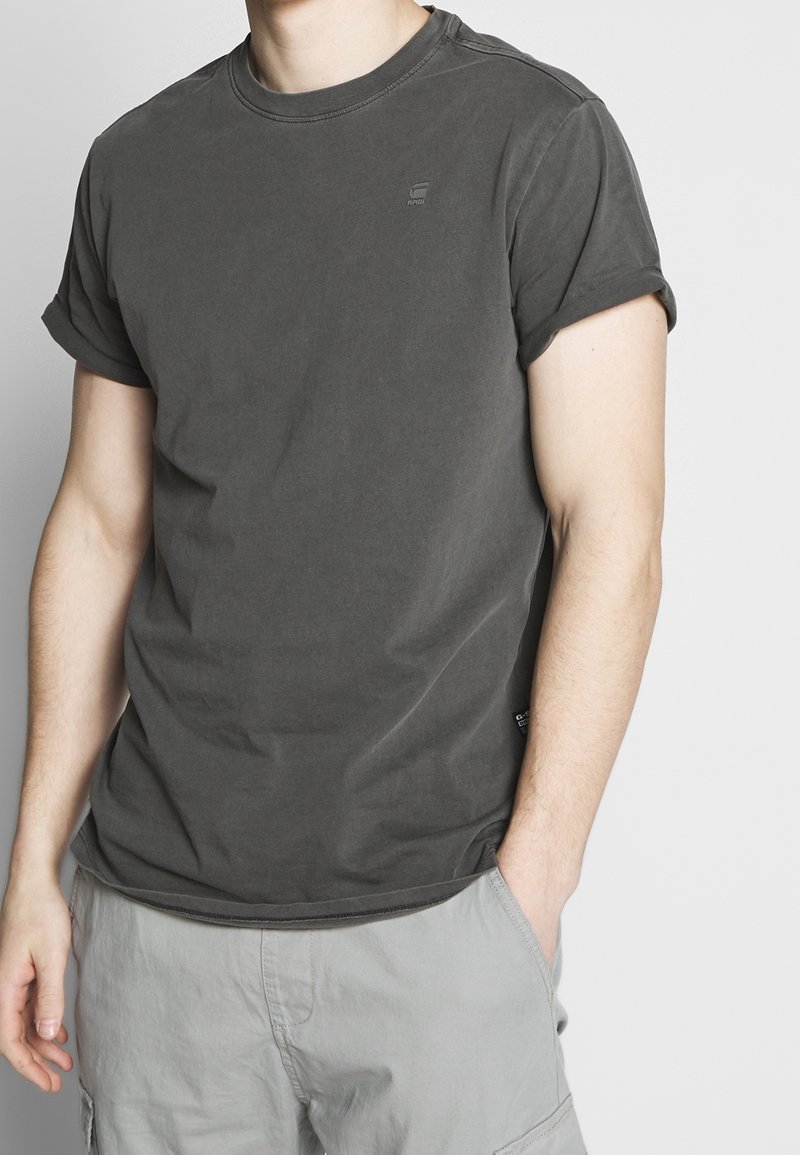 T-shirt en coton gris foncé avec des manches retroussées, col rond et petit logo sur la poitrine. Texture lisse et coupe décontractée.
