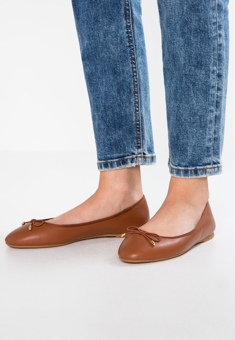 Pieds portant des ballerines en cuir marron avec petits nœuds, associées à un jean en denim bleu avec revers, sur un fond blanc uni.