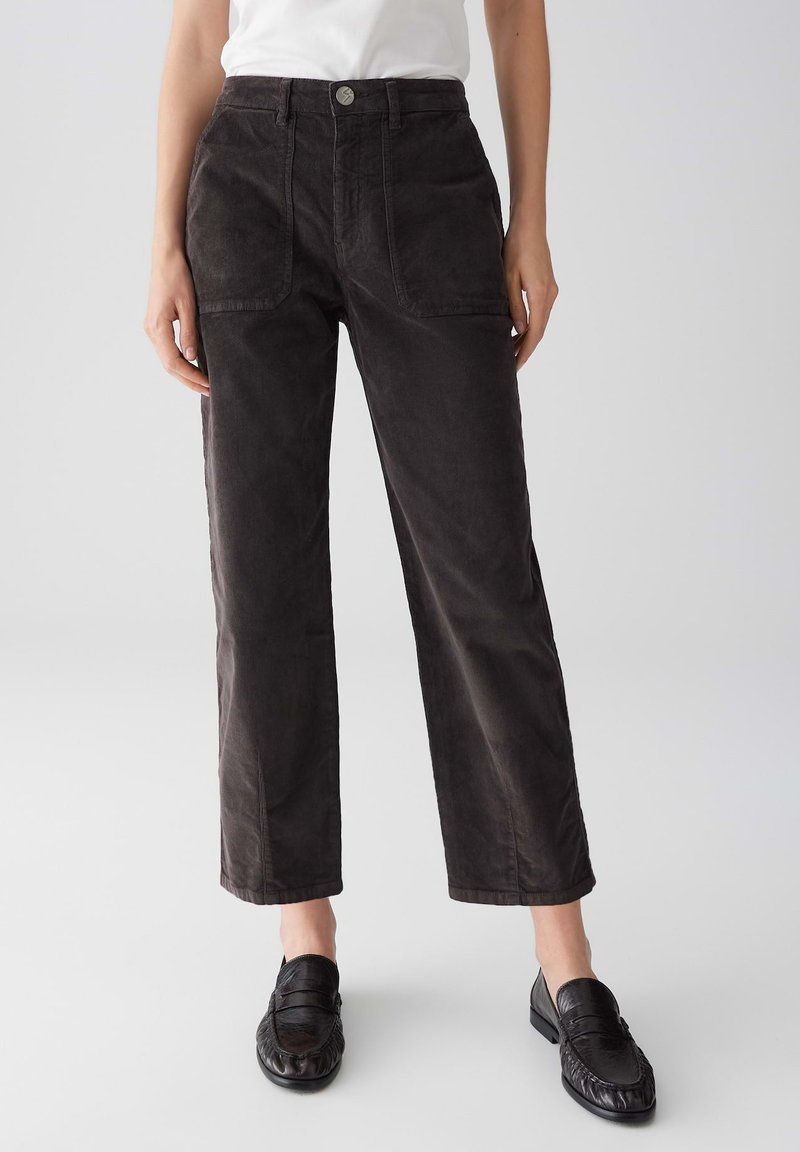 Persona che indossa pantaloni in velluto a coste marrone scuro a vita alta con tasche frontali e mocassini in pelle nera su uno sfondo neutro.