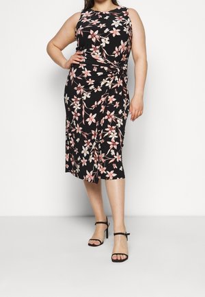 Nainen seisoo käsi lanteilla, päällään musta hihaton midi-mekko, jossa on pinkki ja valkoinen kukkakuosi, sekä mustat korkokengät.