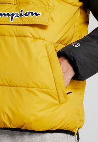 Gul pufferjacka med svarta ärmar, med en frampocket och en logotavla. Slätt, isolerat tyg med sömnadsdetaljer.