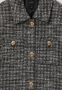 Giacca doppiopetto in tweed bianco e nero, con bottoni dorati e colletto. Tessuto testurizzato con motivo a quadri.