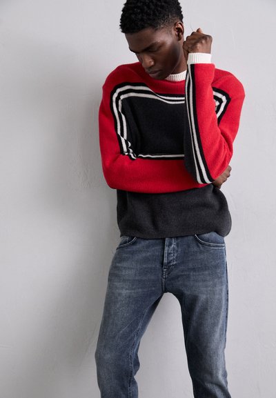 Maglione rosso e nero con strisce bianche, texture a maglia, abbinato a jeans in denim blu. Il maglione presenta spalle scese e polsini a coste.