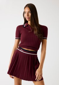 Bordó kötött crop polo, gallérral és bordázott textúrával; párosítva egy matching pliszírozott szoknyával, amely márkázott derékpánttal rendelkezik.