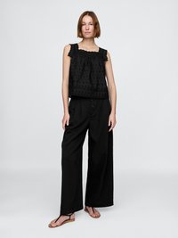 Top senza maniche nero con bordo in pizzo decorativo e texture a pois, abbinato a pantaloni neri a gamba larga e sandali flat.