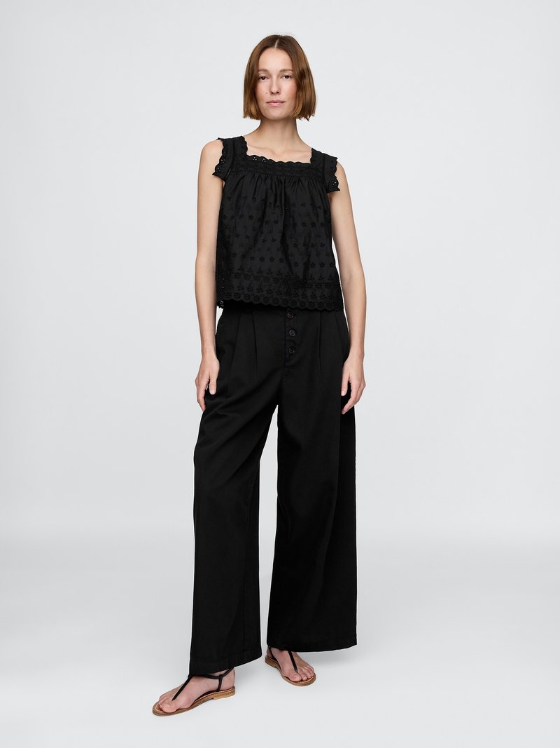 Top senza maniche nero con bordo in pizzo decorativo e texture a pois, abbinato a pantaloni neri a gamba larga e sandali flat.