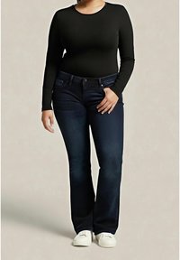 Femme portant un haut noir manches longues, un jean bootcut bleu foncé, des baskets blanches, debout devant un fond beige uni.