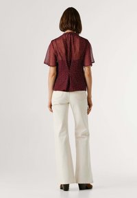 Blouse bordeaux à pois avec manches courtes volantées, fermeture à boutons dans le dos et design fendu ; associée à un pantalon évasé crème et des talons marron.
