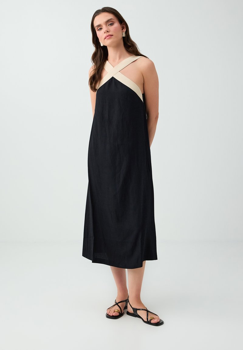 Femme portant une robe midi noire sans manches avec des bretelles croisées beiges et des sandales plates noires, debout devant un fond uni.
