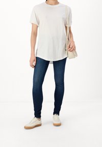 Biała bawełniana koszulka z podwiniętymi rękawami, w połączeniu z ciemnoniebieskimi dopasowanymi dżinsami i białymi trampkami z beżowymi akcentami. Małe logo na koszulce.