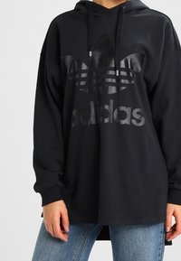 Svart hoodie i bomull med långa ärmar, med en strukturerad Adidas-logotyp och en förlängd design med en kenguruficka.
