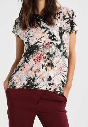 Femme portant un haut à manches courtes à imprimé floral avec des fleurs noires, roses et rouges, associé à un pantalon bordeaux taille haute.