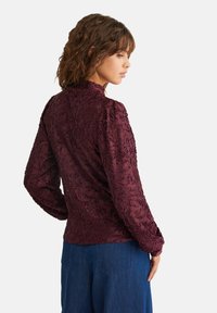 Bluză burgundă cu design floral texturat, guler înalt și mâneci lungi, asortată cu pantaloni din denim albaștri.