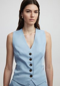 Gilet blu chiaro con scollatura a V profonda, cinque bottoni marroni e tasche laterali. Il tessuto appare liscio con una vestibilità su misura.