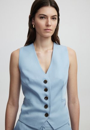 Gilet bleu clair avec un décolleté en V prononcé, cinq boutons marron et des poches latérales. Le tissu semble lisse avec une coupe ajustée.