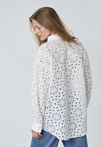 Hvid bomulds skjorte med detaljeret hulsyning, lange ærmer og krave. Bagpå har den en løs pasform og afrundet nederkant.