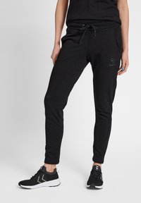 Svarta joggingsbyxor med dragsko i midjan, smalnande ben och sidofickor. Mjuk textur med en diskret logodetalj på låret.