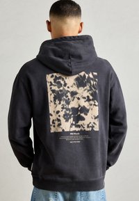 Mand iført sort hoodie med hætten oppe, prydet med et beige og sort abstrakt skovmønster og teksten "Wild Woods" på ryggen, med blå jeans.