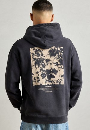 Man wearing black hoodie with hood up, featuring a beige and black abstract forest print and text "Wild Woods" on the back, paired with blue jeans.