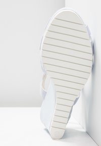 Sandale blanche avec semelle en caoutchouc rainurée, taille 37, positionnée à la verticale contre un mur d'angle blanc et un sol blanc.