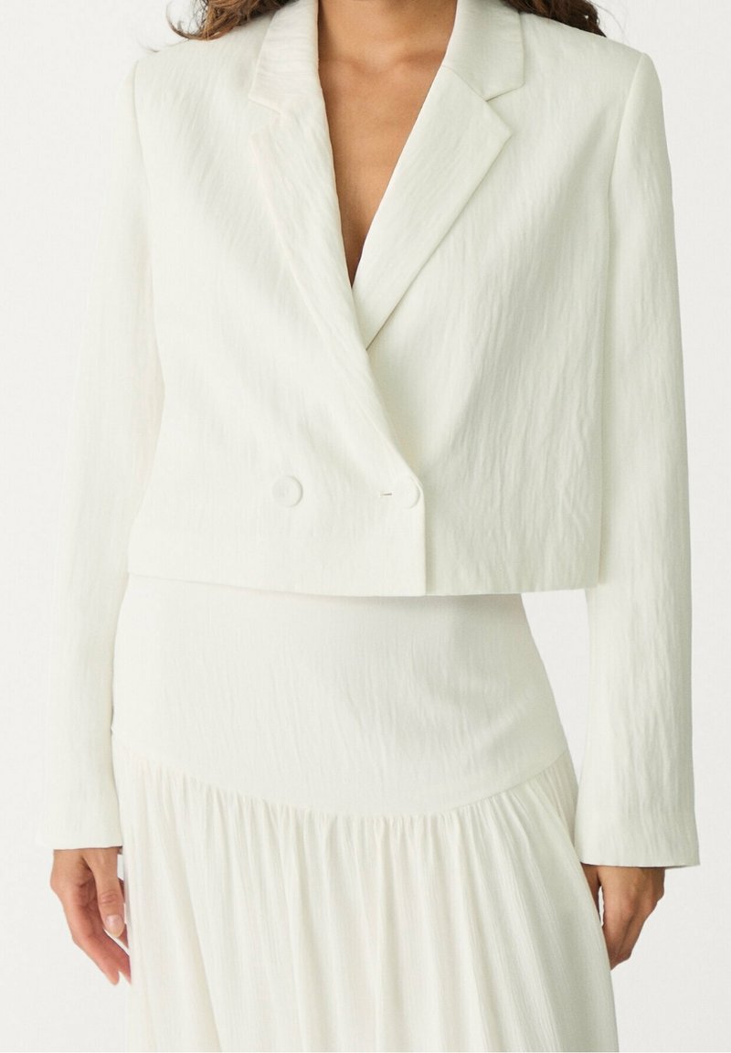 Femme portant un blazer écru texturé croisé court et une jupe taille haute assortie sur un fond uni.