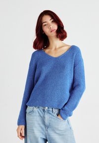 Blå strikket genser med V-hals og ribbestrikket tekstur, kombinert med lyseblå høyhalsede jeans, vist mot en ensfarget bakgrunn.