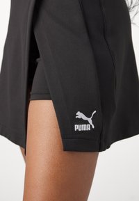 Jupe de sport noire en tissu texturé, avec fentes sur les côtés et un logo Puma blanc sur l'ourlet. Des shorts cachés en dessous sont visibles.