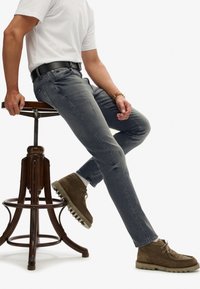Jeans de mezclilla gris con un corte Slim Fit y un desgaste sutil, combinados con botas de ante marrón con cordones y una camiseta de cuello redondo blanca.