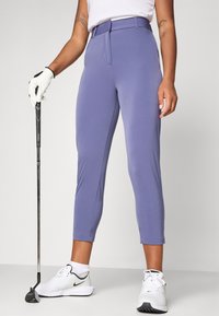 Pantalons de golf violets en tissu extensible, design fuselé avec un pli devant et une coupe longueur cheville, associés à des chaussures de sport blanches.